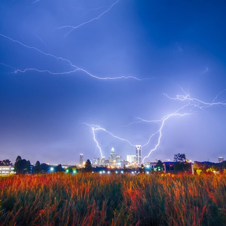 Lightning bolts striking above a city at night with a field in the foreground.